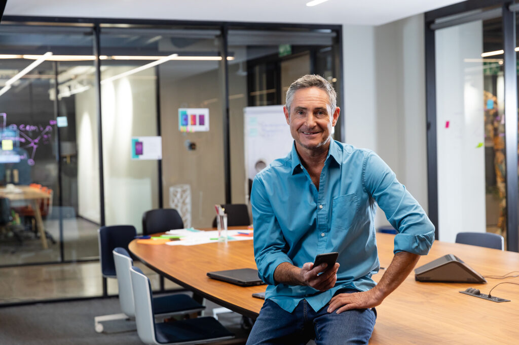 Man sitting on meeting table edge in office meeting room, holding smartphone with laptop and papers. Corporate, modern, glass-walled, executive, business-casual, productivity, brainstorming